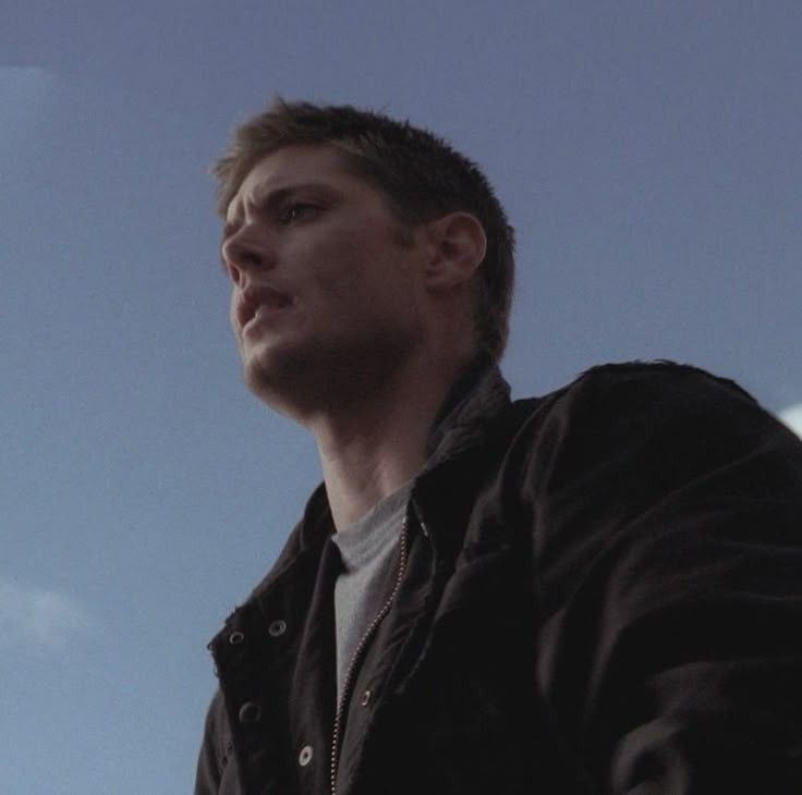 Dean Winchester in a black jacket stands against a clear blue sky, exuding a sense of contemplation or determination in supernatural 