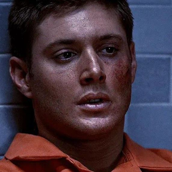 Dean Winchester Jensen Ackles in an orange jumpsuit sits against a blue wall, appearing contemplative in a dimly lit room supernatural 
