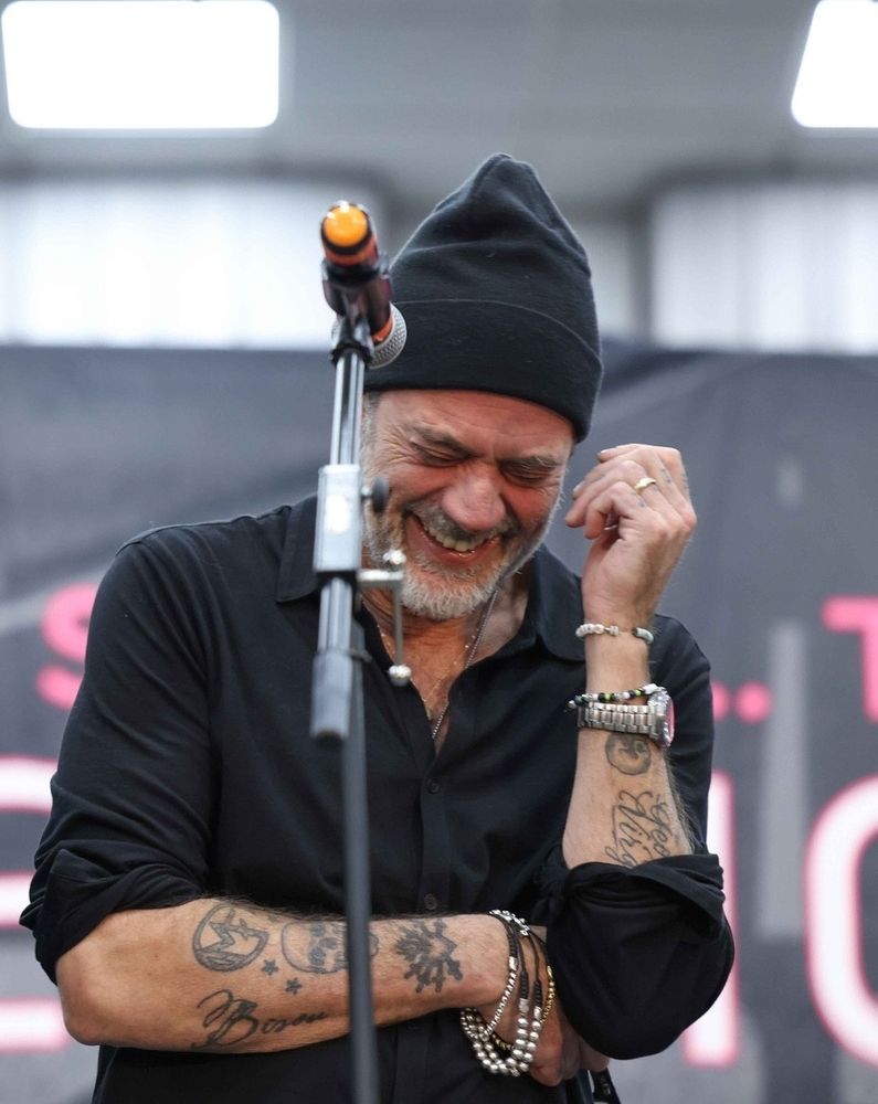 Jeffery Dean Morgan in black shirt with tattooed arms standing in front of a microphone.