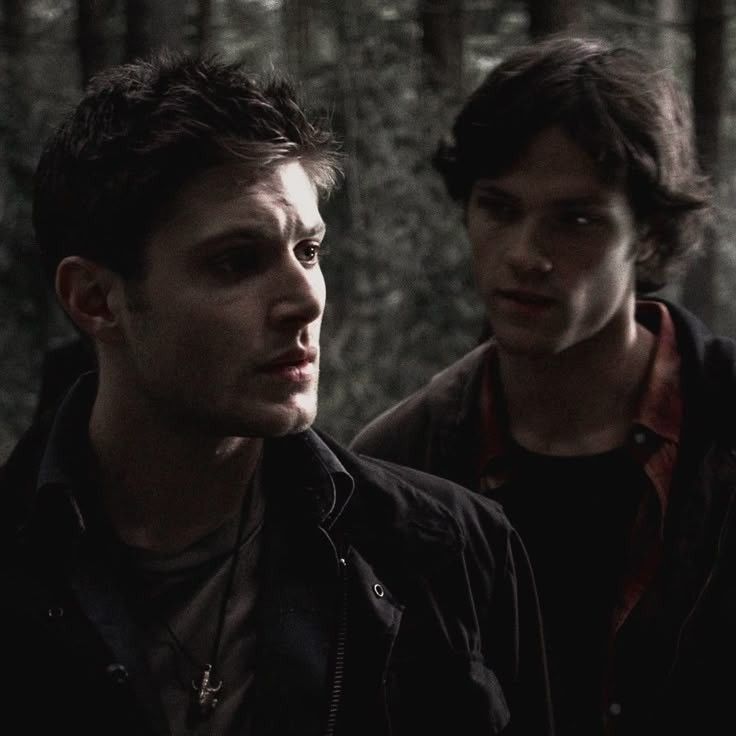 Sam Winchester Jared Padalecki and Dean Winchester Jensen Ackles stand closely together in a dark, dense forest, dressed in casual clothing, exuding a sense of tension and mystery in supernatural 