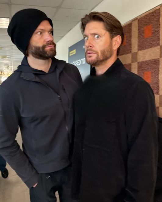 Jared Padalecki and Jensen Ackles are standing close together in a casual indoor setting, wearing dark clothing and pulling weird faces