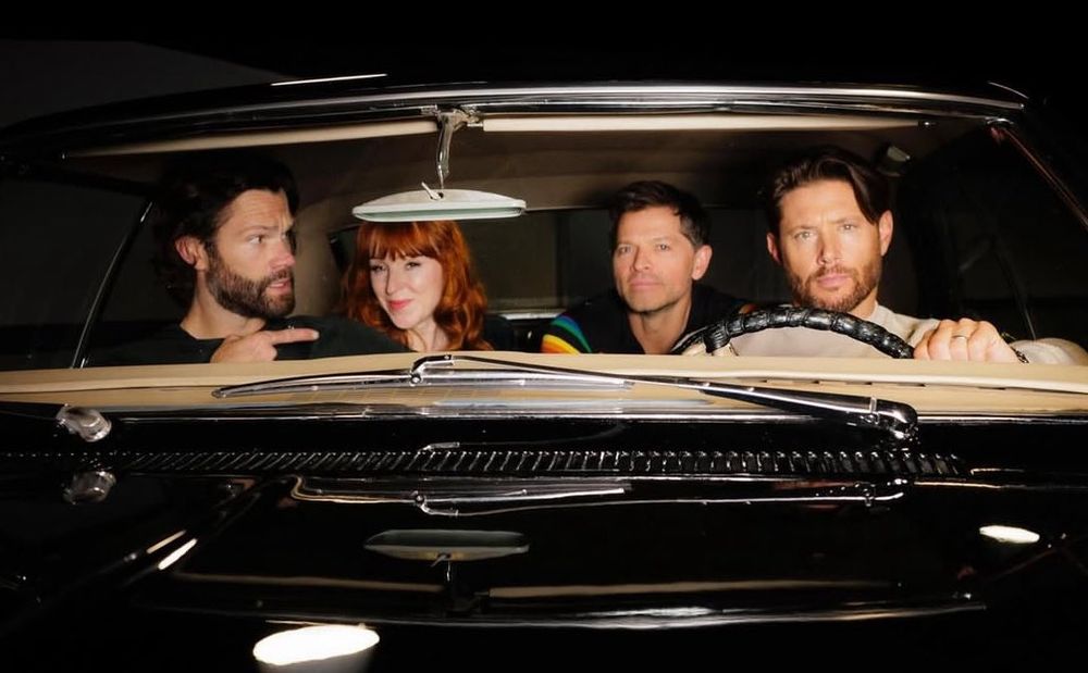 Jared Padalecki Jensen Ackles Ruth connell and Misha Collins sit inside baby, with the dashboard and steering wheel visible in a dimly lit setting.