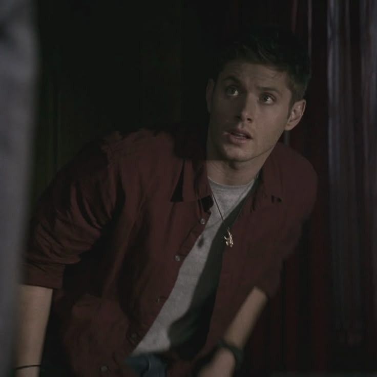 Dean Winchester Jensen Ackles in a maroon shirt leans slightly forward, with a dimly lit background featuring red curtains and shadowy details in Supernatural 