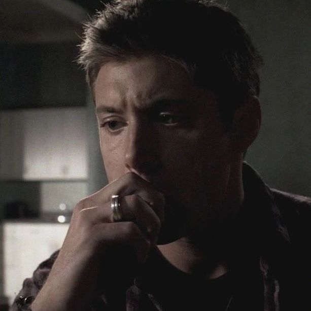 Dean Winchester (Jensen Ackles) sits in a dimly lit room, thoughtfully resting their chin on their hand, with a blurred background suggesting a kitchen in supernatural 