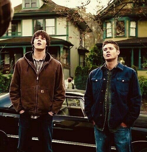 Sam Winchester and Dean Winchester (Jared Padalecki and Jensen Ackles) casually in front of a vintage car, with charming homes in the background, showcasing a cool, autumn vibe.