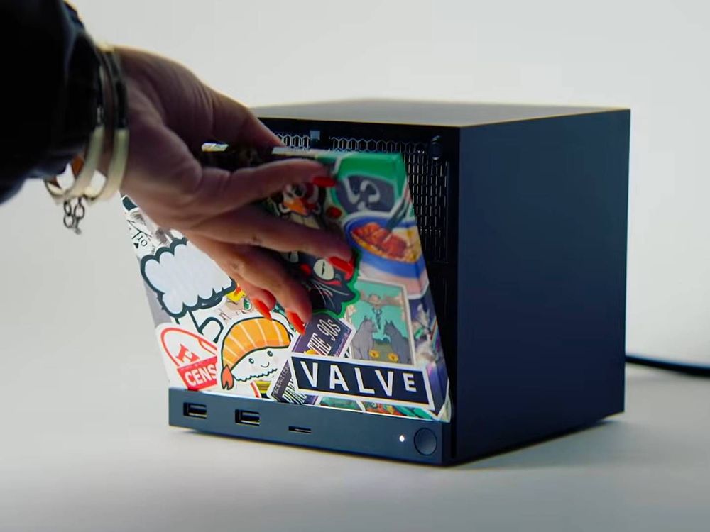 A close-up photo of a person putting a faceplate onto the front of Valve's new Steam Machine. The person's hand has red polish on the fingernails, and a gold bracelet on the wrist. The faceplate is designed in the "Sticker bomb" style, as if it's covered with multiple layers of colorful stickers. The Valve logo is on the bottom right corner of the faceplate.