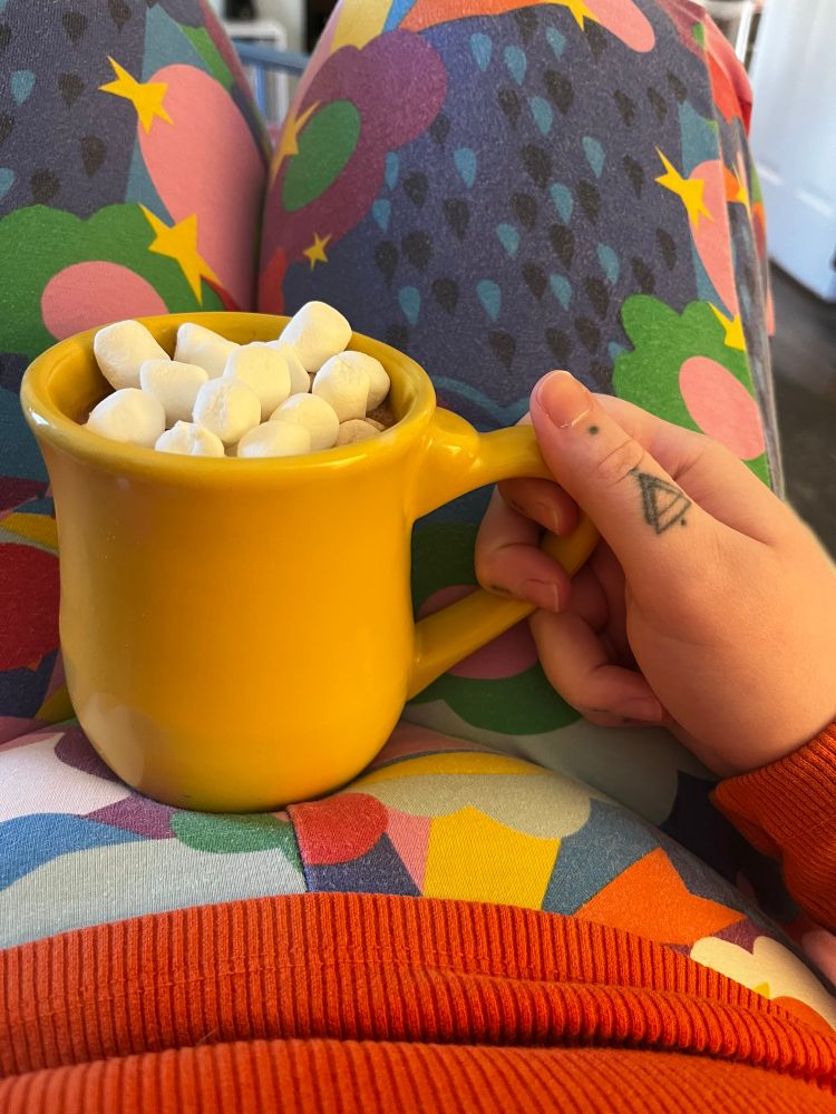 Harper holds a bright yellow, heavy mug full of cocoa and marshmallows on their lap.