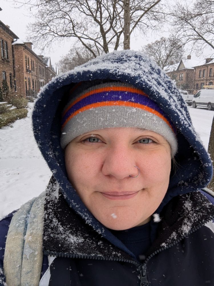 Selfie of me outside on a snow covered street. I'm wearing a hat under a hoodie. The hood is covered in snow. My eyes are crinkled at the corners and there is a small smile on my face.