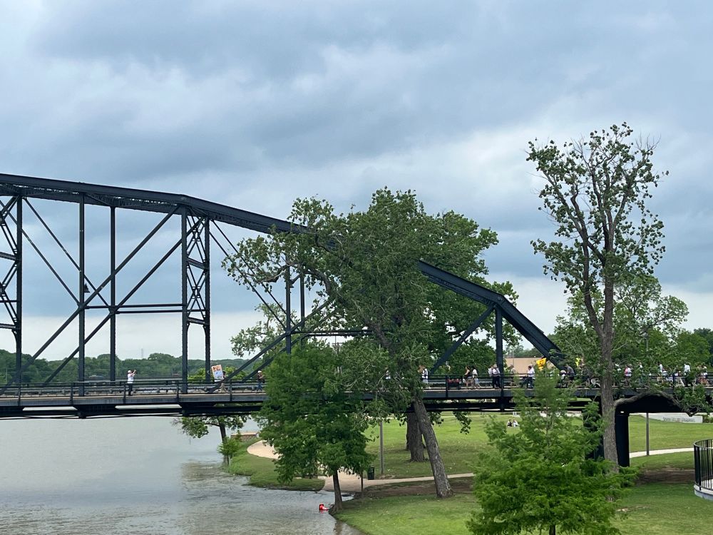Image of a suspension bridge going from the land out over a river. There are people walking along the sidewalk to cross the bridge carrying their protest signs.