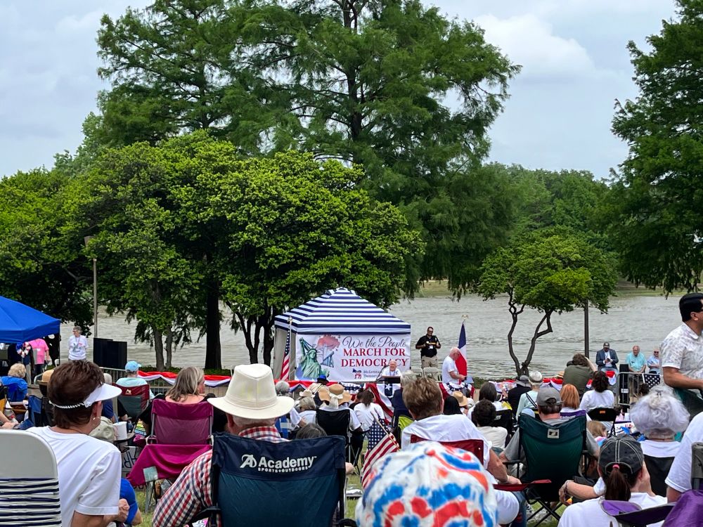 In front of a river, under a big tree, there is a tent and a dais. There is a man at the microphone and a crowd sitting in lawn chairs is listening and cheering. 