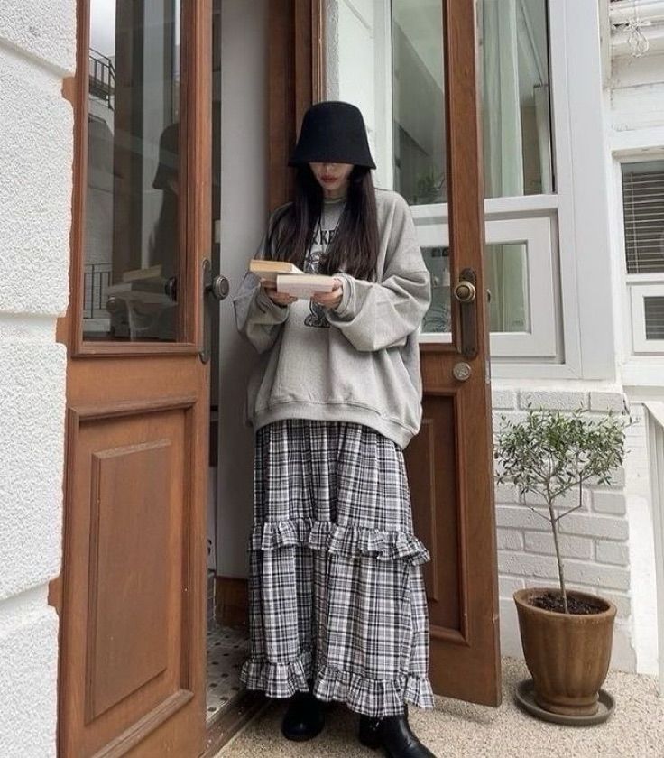 full body photo of a girl with long brown/black hair wearing a black bucket hat covering half of her face. she’s wearing an oversized gray hoodie with some sort of graphic mostly covered by her arms holding two books in front of her; and a gray and white plaid maxi skirt with ruffles in both the bottom and the middle of the skirt, and finally some black boots.