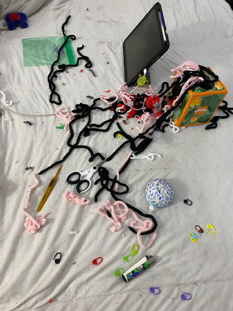 A bedspread covered in stitch markers, crochet hook, scissors, yarn scraps, lighter, iPad, and ziploc bag