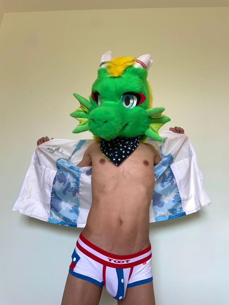 An adult male wearing a green dragon fursuit head, dark blue bandana, a blue and white jacket, and underpants made of white, red and blue garments, standing and making an undressing pose to open up his jacket, revealing his skinny body and an underwear bulge made eye-catching by his half erect penis.