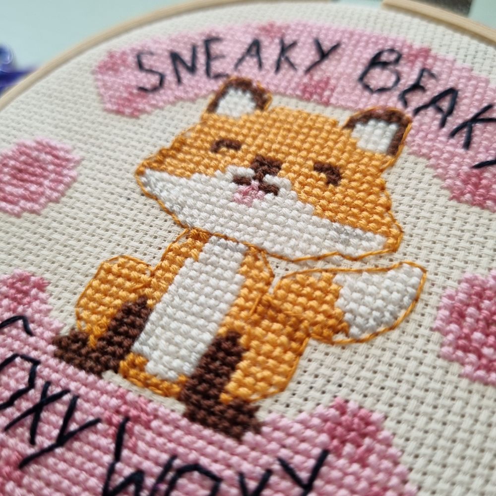 Close up of a cross stitched fox, orange with brown paws and ears. There's pink banners in the background and text that says "sneaky beaky foxy woxy"

Other text variations not shown: "Please [fox] off" and "I've got no [fox] to give".