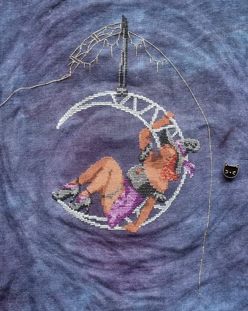 Cross stitch of a witch wearing black and purple, sitting in a moon lyra