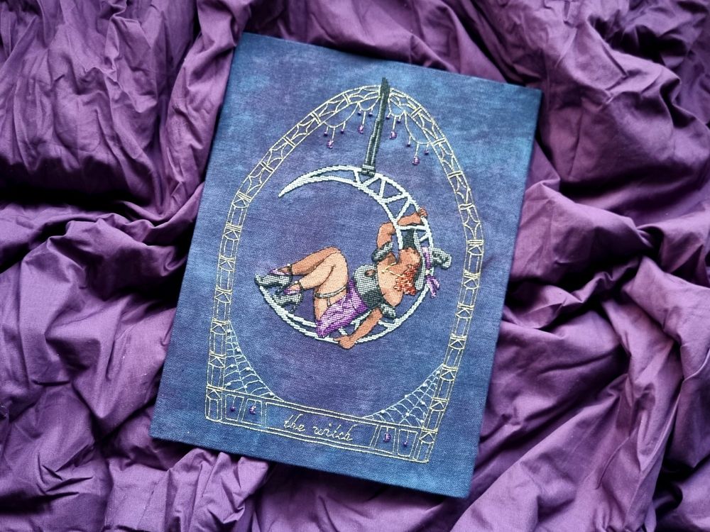 Cross stitch of a witch sitting in a moon lyra, surrounded by a golden border. Text at the bottom says "the witch".