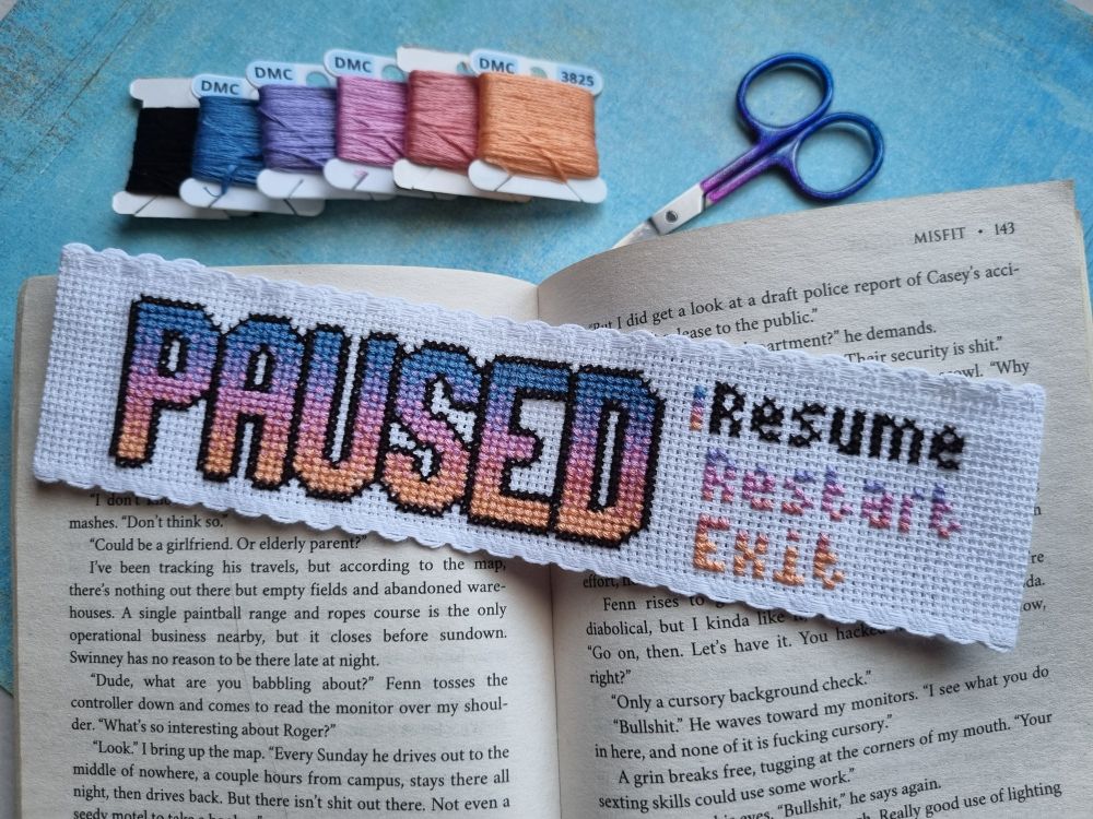 Cross stitched bookmark with the word "PAUSED" in all caps, and "Resume", "Restart", and "Exit" beside it.
Bookmark is resting on an open book, with bobbins and scissors in the background