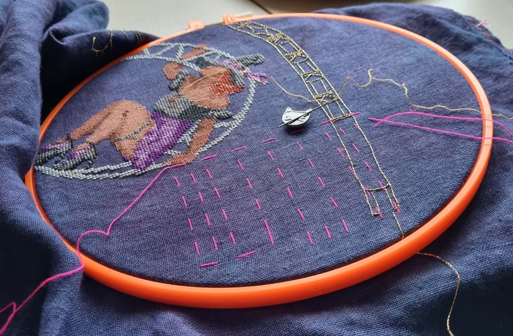 Cross stitch of a girl in a moon lyra, with pink thread creating the start of a grid in the bottom right corner