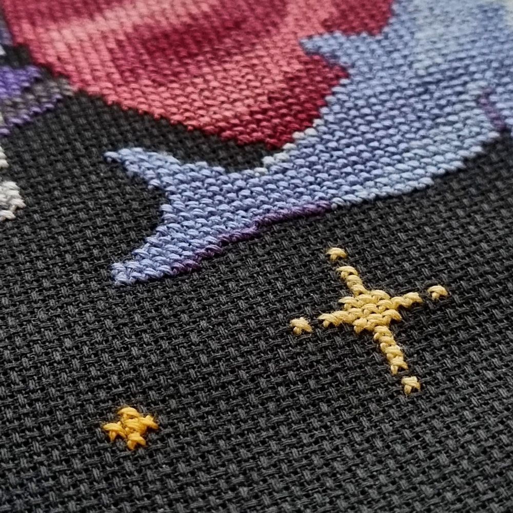 Close up of cross stitch on black aida showing orange/yellow stars, and a blue/purple tail.