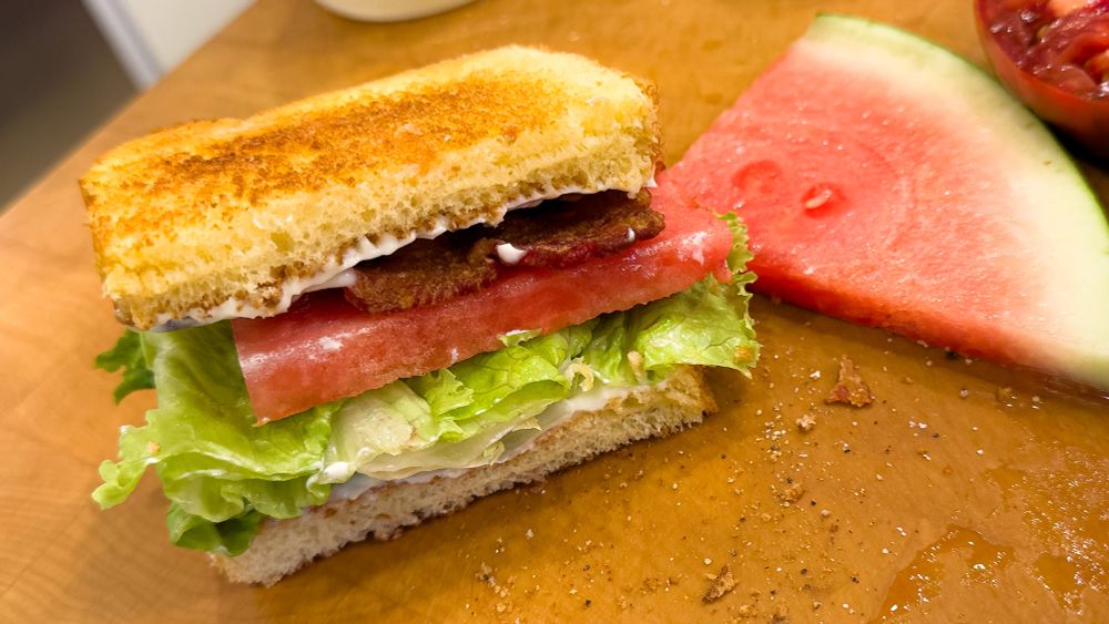 A sliced BLT sandwich sitting next to a watermelon wedge on a wooden cutting board, but in place of the slab of tomato, there is a slice of watermelon instead.