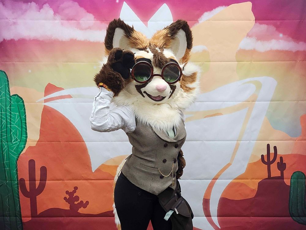 A tan, brown, and white cat partial fursuiter in a white button down shirt, a houndstooth vest with a pocket watch chain, a light green tie and black pants posing in front of a western backdrop. One of their paws holds their glasses.
