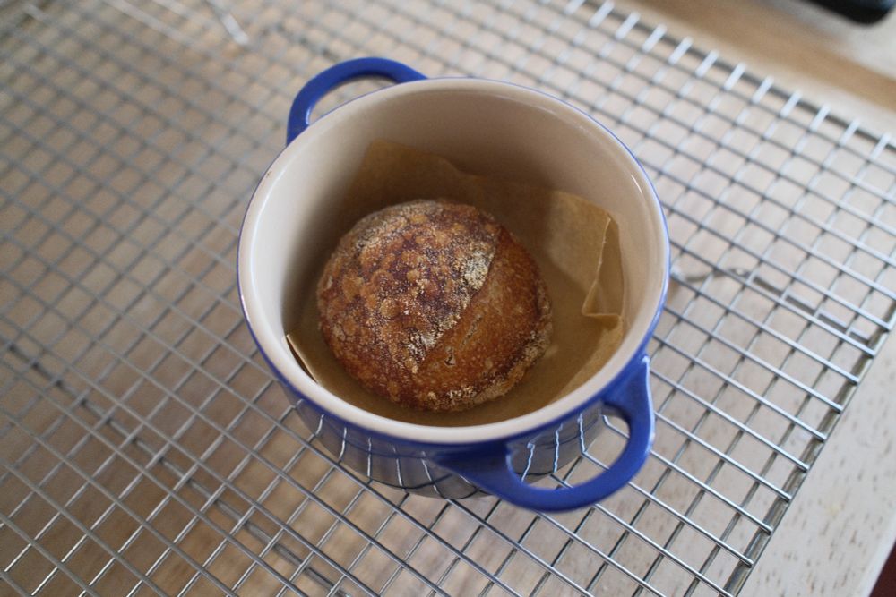 An overhead photo of a small artisan style bread boule on a small scrap of parchment paper inside an equally small ramekin that looks like a tiny le creuset dutch oven. The appearance is exactly like a normal sized bread boule in color and scoring but is overall about the size of a golf ball.