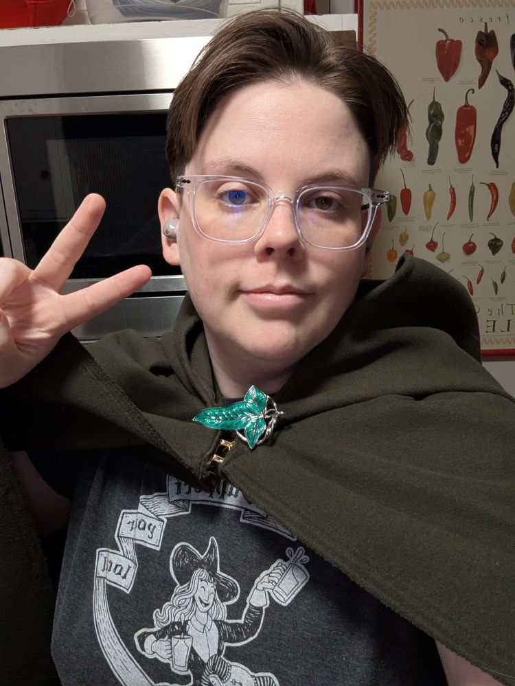 Katie is facing the camera holding a peace sign while wearing a dark green hooded cloak held together at the neck with a leaf brooch in the style of those seen in Lord of the Rings.