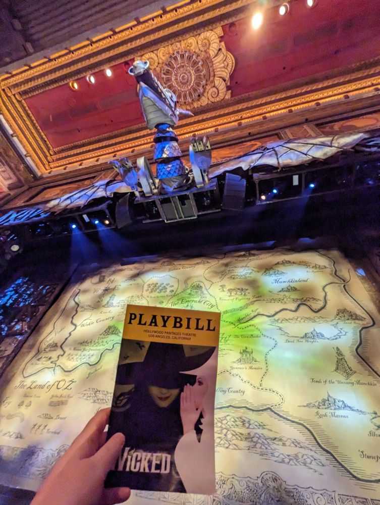 A hand is holding a playbill for the musical Wicked, the background is the stage curtain which shows a map of the land of Oz with a mechanical dragon above it.