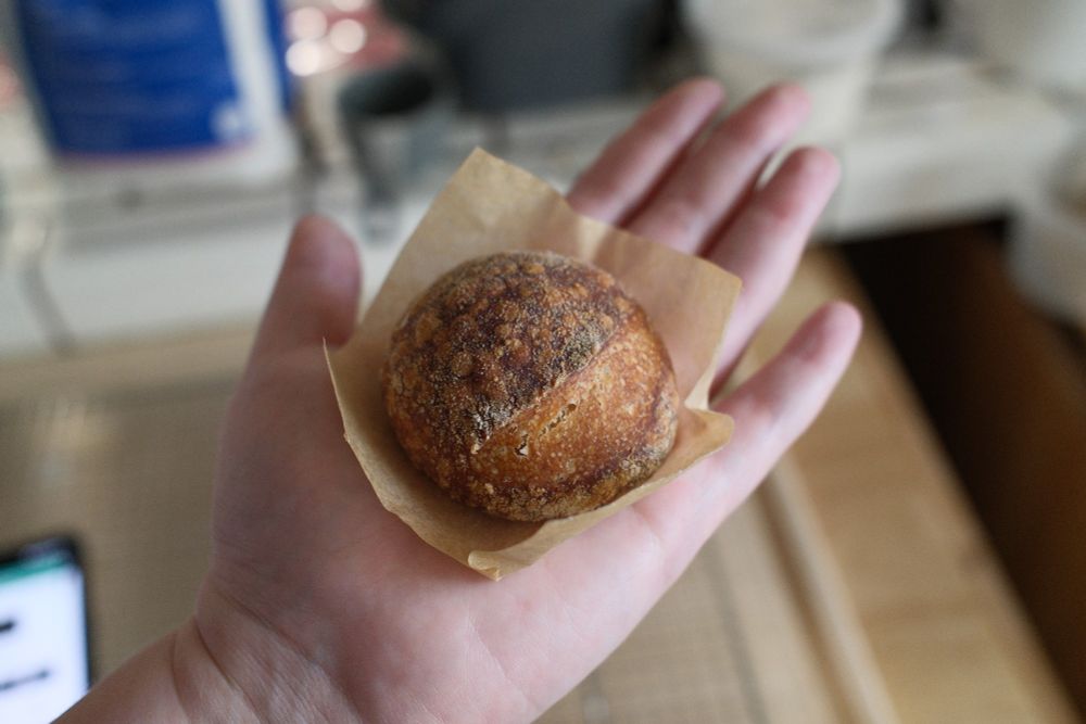 A hand holds the small bread boule and parchment paper, the bread is small enough to fit inside the palm of the hand.