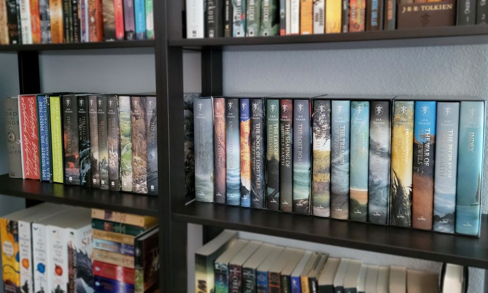 Closeup of two adjacent bookshelves with a connecting row of Tolkien books with oh so pretty matching dust jackets. All of the colorful illustrated covers are matte, with the titles standing out in shiny gold foil. On one end of the row, three books are displayed backwards, with the spines towards the wall, to show off the colorful sprayed edges with white text written in the Cirth and Tengwar scripts invented by Tolkien.