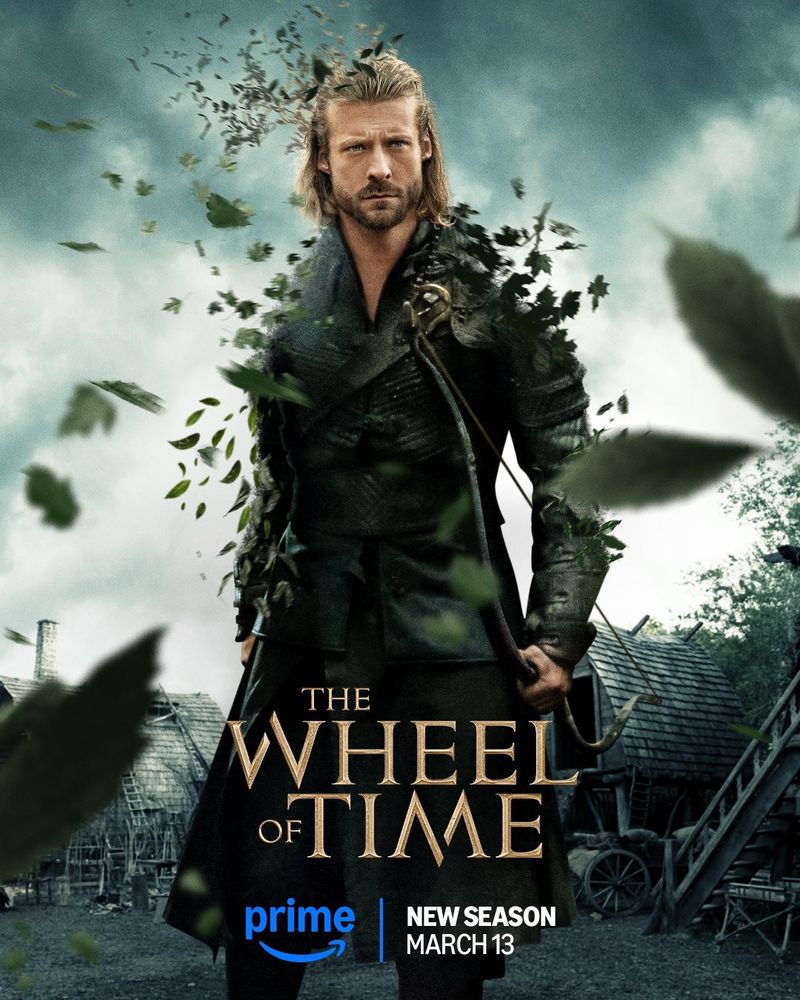 A poster of Maksim (Taylor Napier) in The Wheel of Time Season 3. He stands in a village, the sky looking stormy, wearing a sturdy black leather coat and holding a short bow in his left hand, give a stoic blue steel expression. his body and armour are beginning to dissolve into swirling leaves. The Wheel of Time logo is center at the bottom of the image, with the Prime Video logo beneath and text reading: “New Season March 13”.