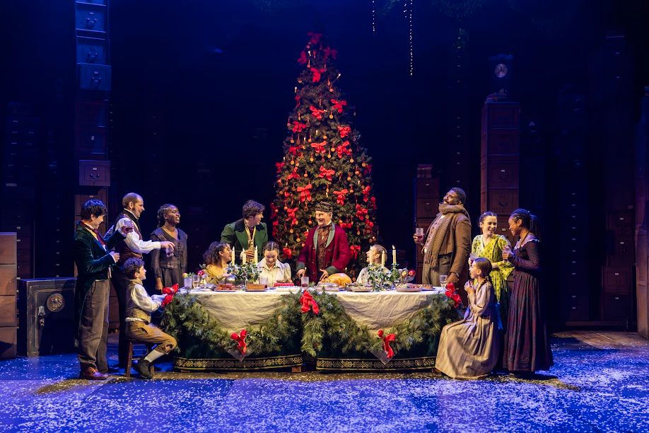 Production photo shows the cast sat around a table for Christmas, behind them is a Christmas tree