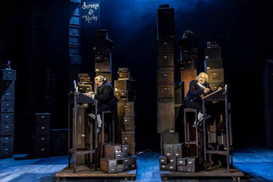Production photo shows Scrooge and Marley working at high desks behind which are piled high filing cabinets
