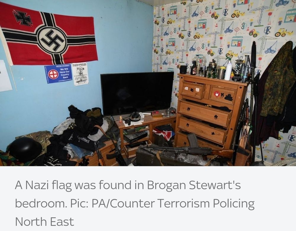 Picture of a messy bedroom with a flag including a swastika and an iron cross. Next to this wall is another wall with wallpaper with childish wallpaper including cranes and diggers.