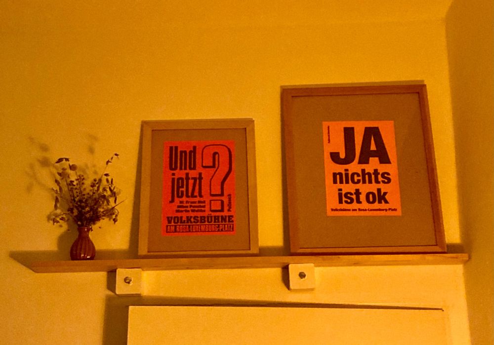 Neonrotes Poster von „Und jetzt“ und neonoranges Poster von „Ja nichts ist ok“ in einfachen Holzrahmen auf einfachem Regalbrett. Daneben Trockenblumen.