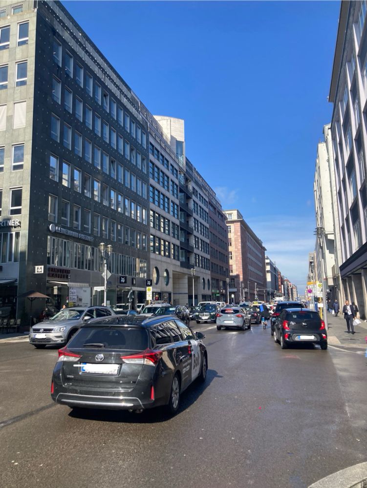 Viel befahrene Straße, pro Richtung eine Spur, rechts und links parkende PKW. Eingekeilt zwischen fahrenden und parkenden PKW eine Person auf Fahrrad
