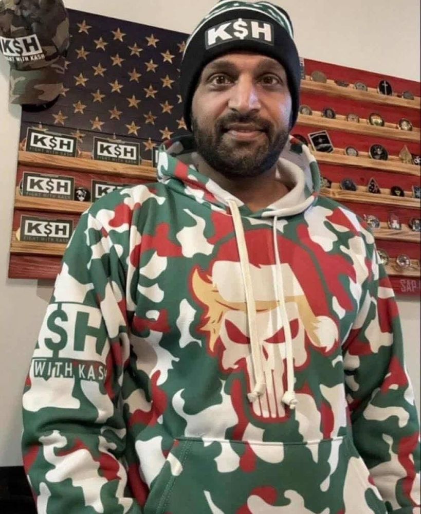 Kash Patel wearing the corniest Christmas camo punisher skull sweatshirt with his own podcast advertised on the sleeve, wearing his own merch hat too and another behind him, in front of an American flag challenge coin holder that also contains stickers for his podcast 