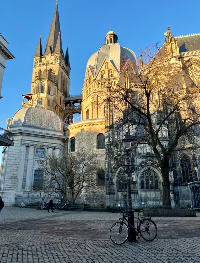 Aufnahme der Südseite des Aachener Doms. Turm, Kuppel und das Dach des Kirchenschiffs sind von der Sonne beschienen und in goldschimmerndes Licht getaucht. Darunter liegt das Gebäude im Schatten. Durch ein Kirchenfenster in der Mitte ist ein kleiner Lichtfleck zu erkennen. Der Himmel über dem Dom ist hellblau und wolkenlos. Im Vordergrund hebt sich dunkel ein blattloser Baum ab, davor steht eine Straßenlaterne, an der ein Fahrrad lehnt.
