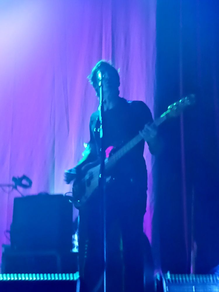 Bass player for Manchester Orchestra. He is rocking out and playing the bass with ultimate coolness. There is a purple curtain in the background. 