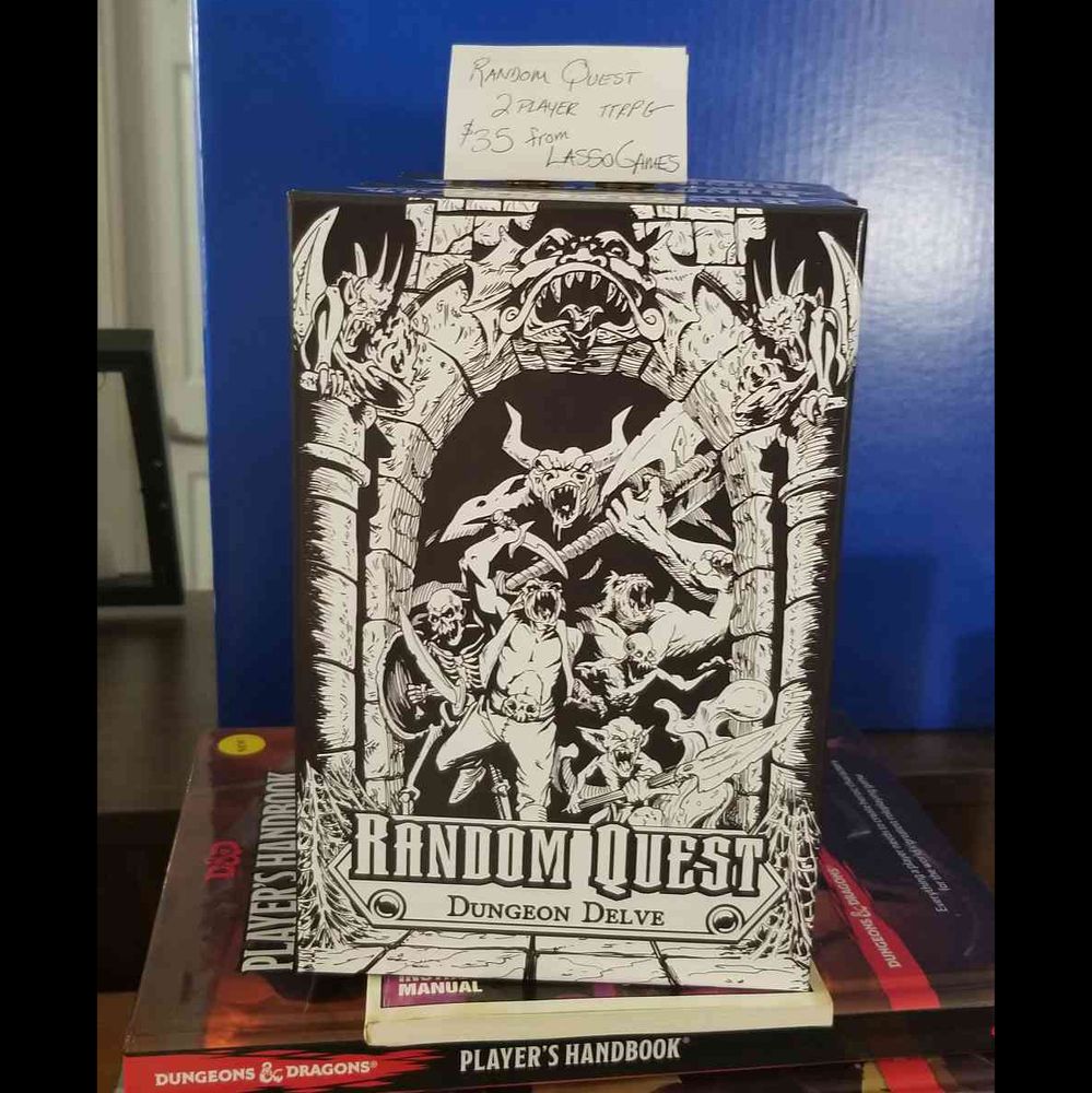 a image of Random Quest box sat upright on a few copies of dnd's player handbook. A label is on top that reads "Random Quest 2 player ttrpg 35$ from LassoGames!