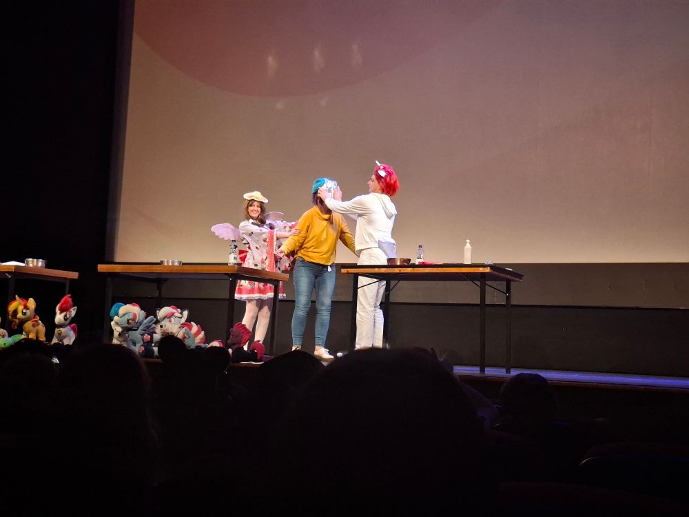 3 people are standing on a theater stage, wearing My Little Pony themed cosplay. The person on the right is pressing a cream pie in the face of the center person while the third person is shoving them towards the pie.