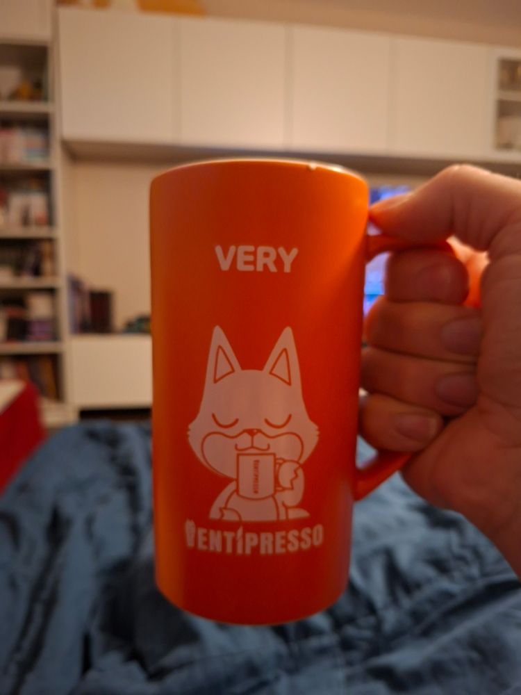 Photo shows a hand holding a tall orange coffee mug from korean coffee shop Ventipresso. On it there's a white print showing a korean style comic fox enjoying a cup of coffee with his eyes closed. It looks like they'd inhale the smell of the coffee. Over them it reads 'VERY', which is the name of the Ventipresso fox mascot.
