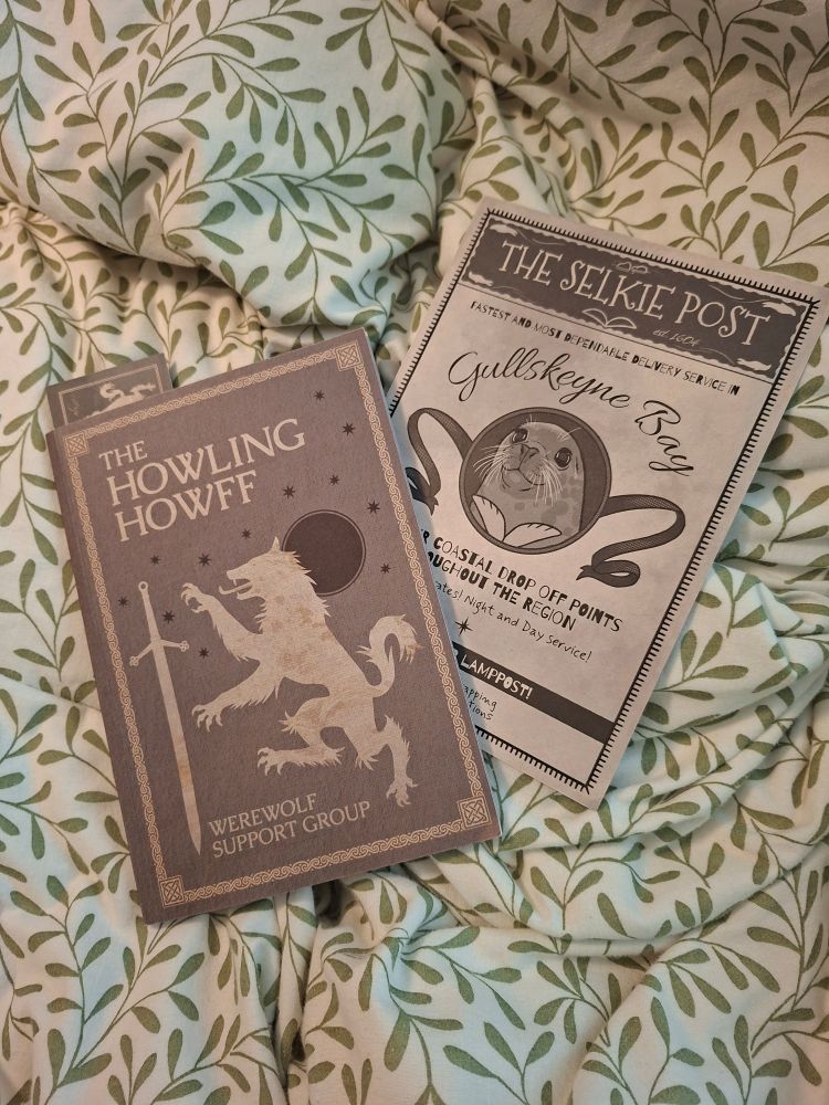 Photography shows a greyscale booklet and a flyer. The Book reads 'The Howling Howff - Werewolf Support Group' and shows a stylused Werewolf next to what appears to be a Claymore sword with a dark full moon behind the wolf.
The flyer shows a Seal (or Selkie in this case) and appears to advertise the Gullskeyne Bay Selkie Post postal service.