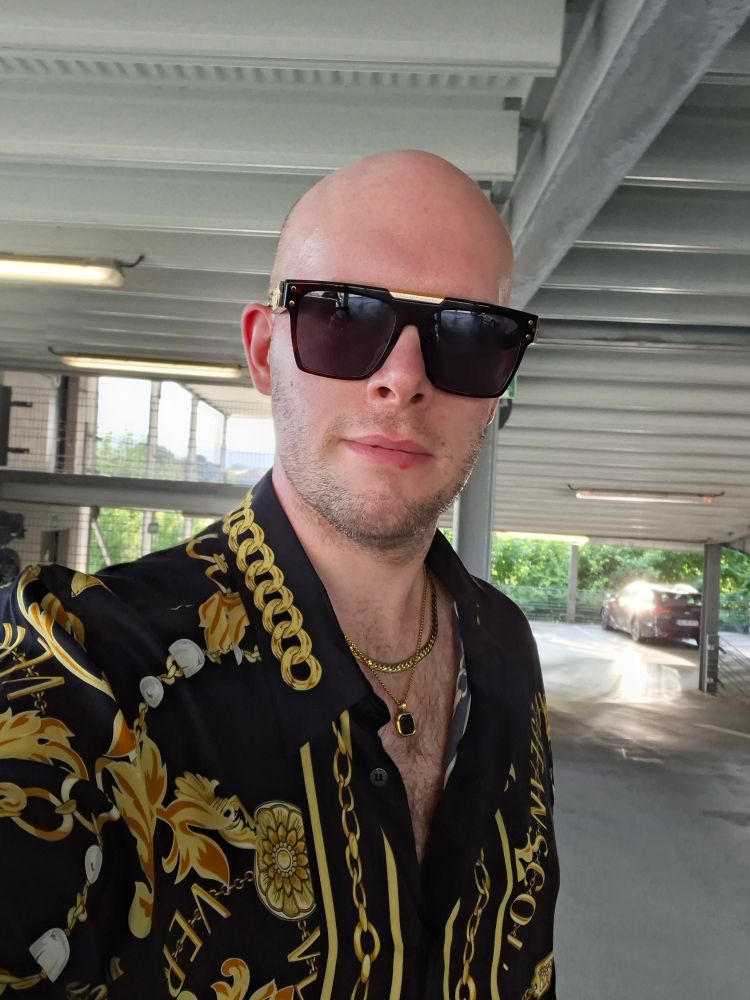 Selfie shows a bald white man in his 30's. He's got a short 3-Days beard and wears a black Versace shirt with golden chain print. He also wears a gold colored double necklace and large dark sun glasses by Phillip Plein.
