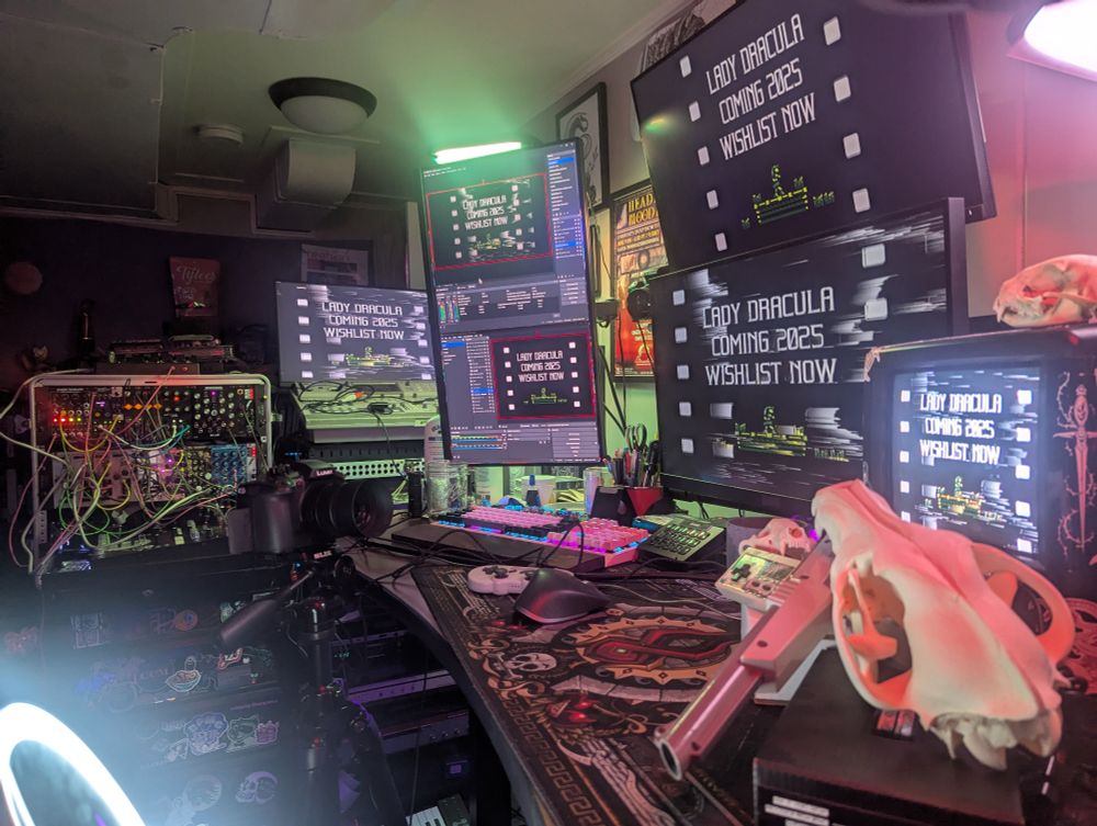 Multiple screens displaying the text 
"LADY DRACULA
COMING 2025
WISHLIST NOW"
Some are glitchier than others. My analog rig is in the background. OBS is open on multiple screens. There's a camera. And skulls.