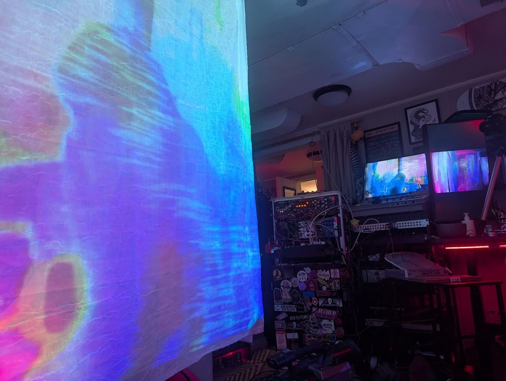 Photo of Zoë's studio - a glitchy silhouette is projected onto a sheet in the foreground. In the background, a wall of glitch gear is visible.