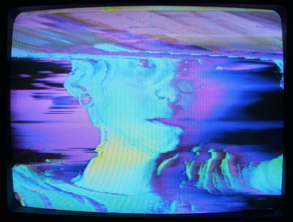 Zoë's face on a CRT screen - very blue, very glitchy. She is surrounded in a purple haze.
