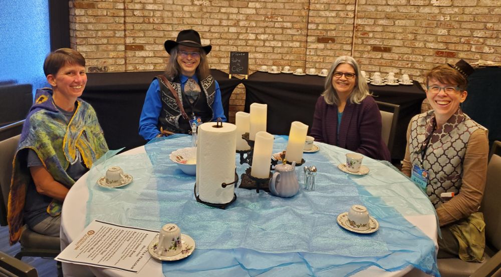 Clara Ward, Anthony Francis, Betsy Miller, and Liza Olmsted are sitting at a round table having tea during Clockwork Alchemy. All are smiling