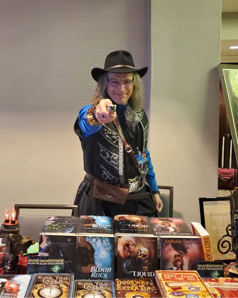 Anthony Francis is standing behind a table with stacks of his books on it. He is wearing a black cowboy hat with a black and silver vest, and aiming a sonic screwdriver at the camera.
