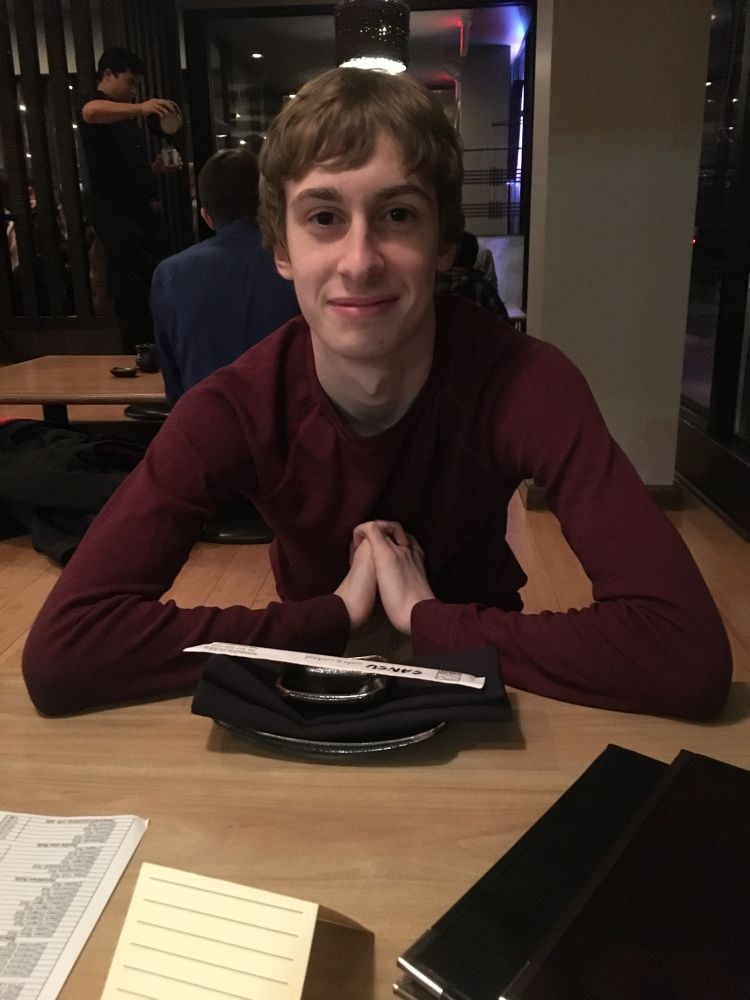 Photo of 20yo pre-transition Luna at a sushi restaurant. She has a short haircut and a slight smirking smile. Her hands are folded together in front of her as she waits patiently for her food.